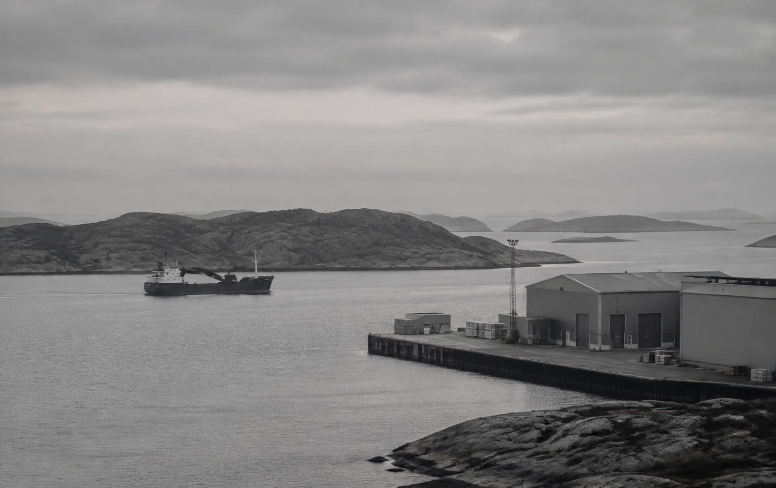 Small cargo vessel approaching a Norwegian coastal quay and warehouse under flat morning cloud