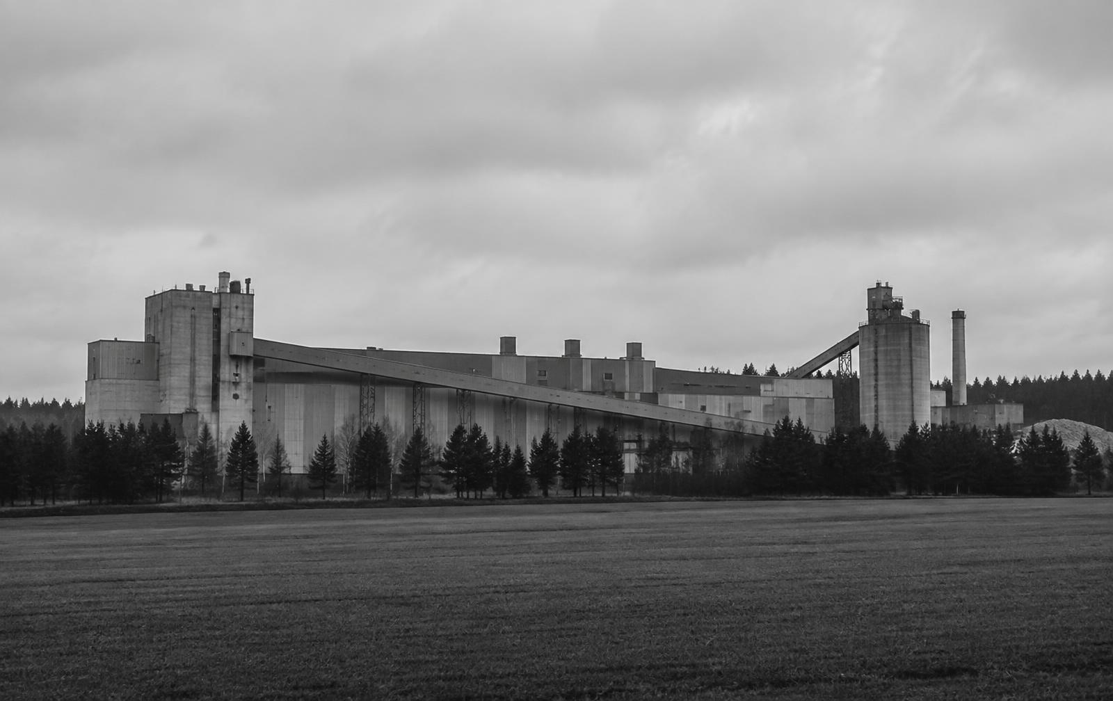 Long industrial plant facade beside flat farmland and a pine treeline under overcast Swedish light